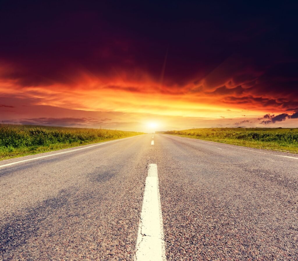 A long, empty asphalt road stretches toward a glowing sunset over a vast, grassy field under a drama…