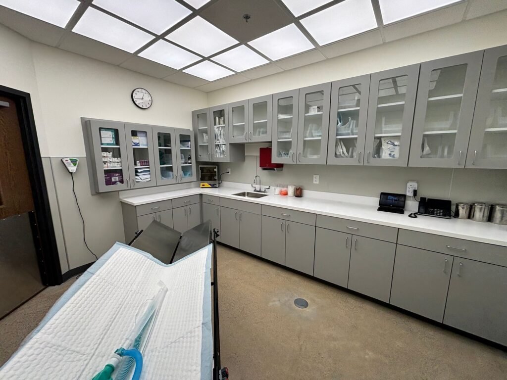 A clean, modern medical examination room featuring gray cabinetry, a sink, and a paper-covered exam…